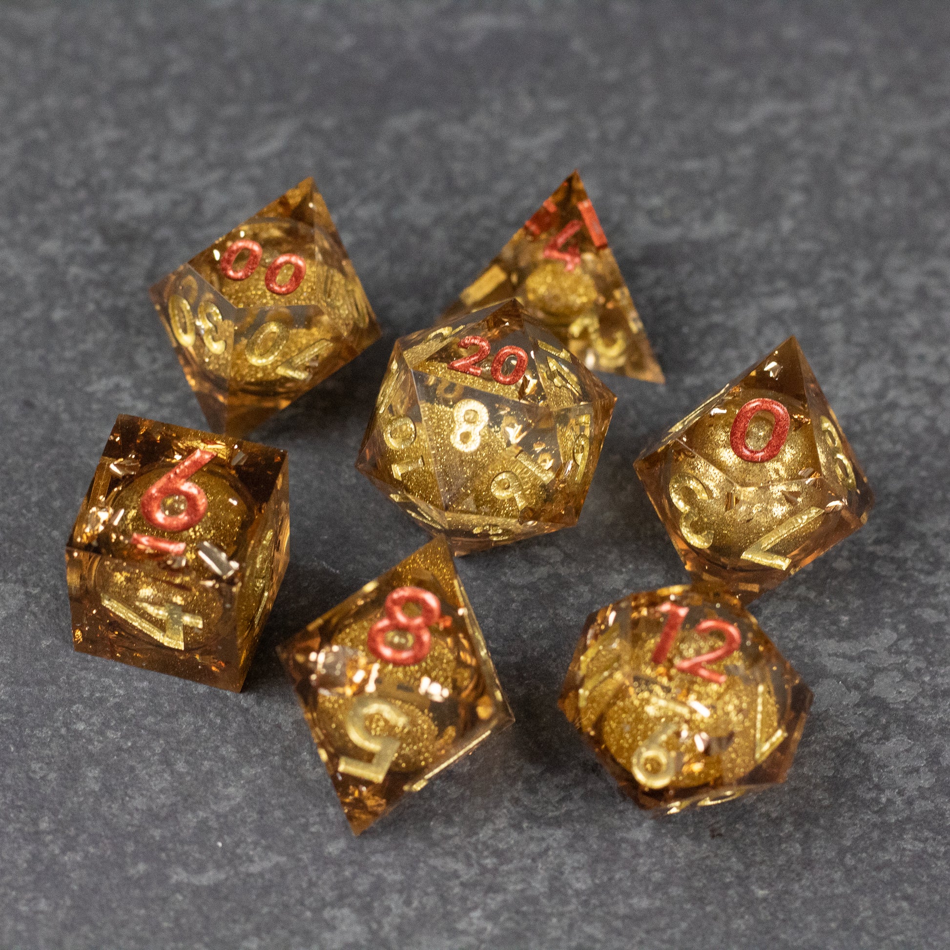 Dragon's Hoard Liquid Core Dice Set - Seven gold translucent polyhedral dice with sharp edges and red numbers on a dark gray surface.