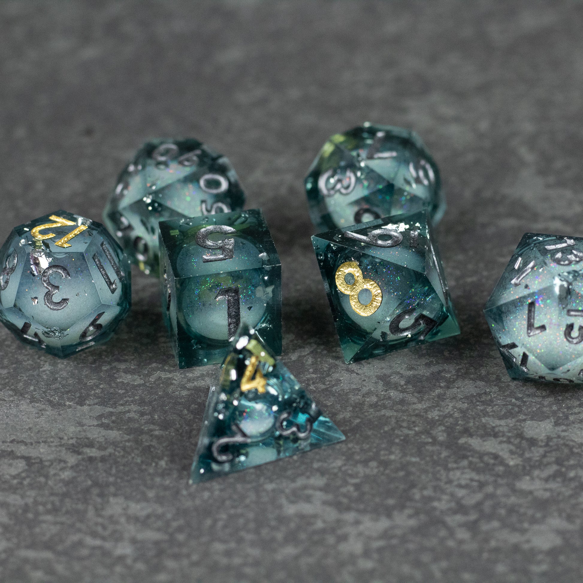 Call Of The Deep - Liquid Core Dice Set - Translucent teal liquid core polyhedral dice set with sharp edges and gold numbering on gray surface.