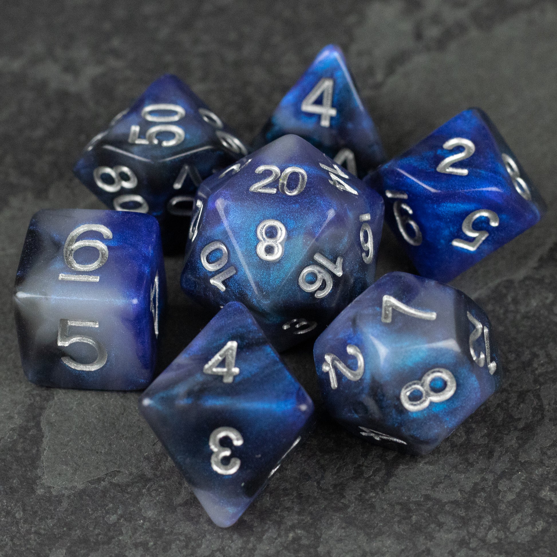 Cosmic Dust Dice Set - Blue and purple marbled polyhedral dice set with white numbering on dark stone surface.