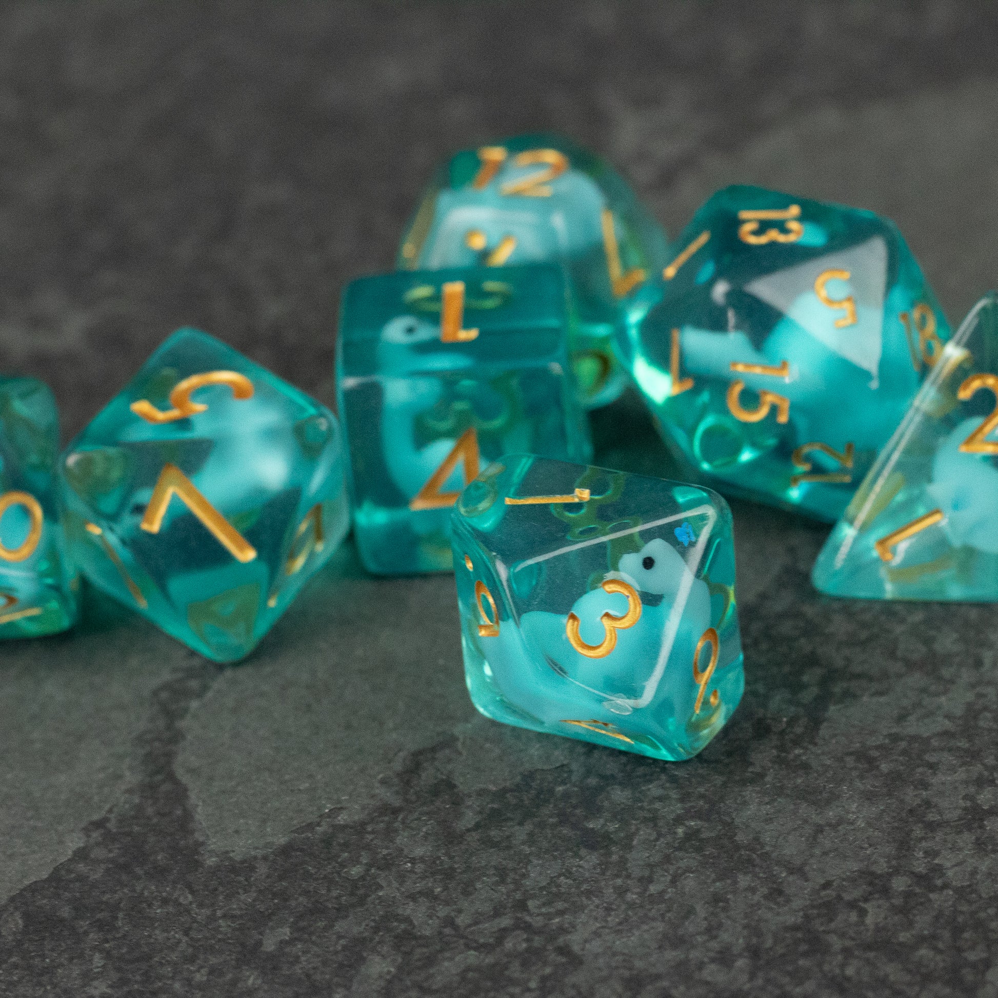 Blue Brontosaurus Dice Set - Translucent blue polyhedral dice set with gold numbers, including d20, d12, d10, d8, d6, and d4 on dark surface.