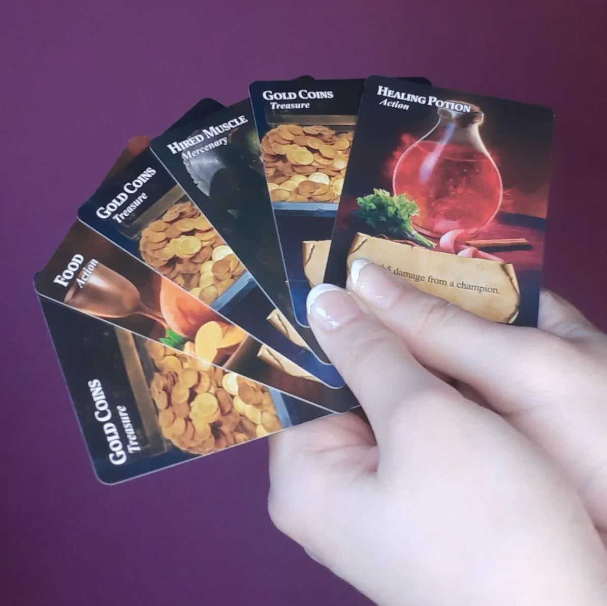 Dragonvault: Original Game - Hand holding five Dragonvault game cards with potion and coin illustrations on purple background.